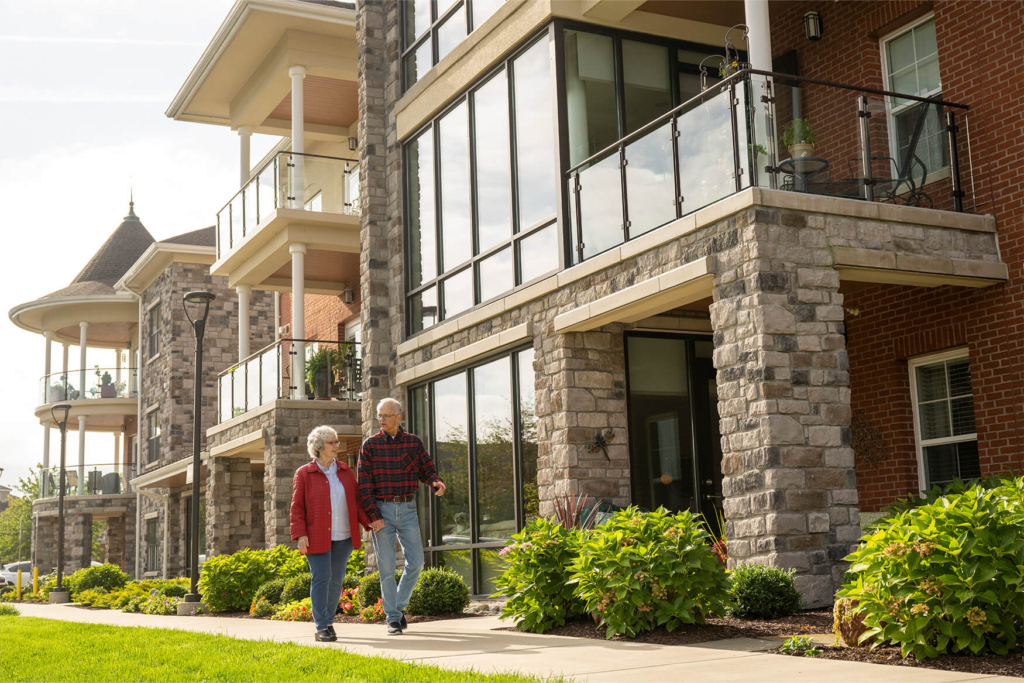 A couple walks down the sidewalk outside an apartment building on their 1-bedroom senior apartment living tour