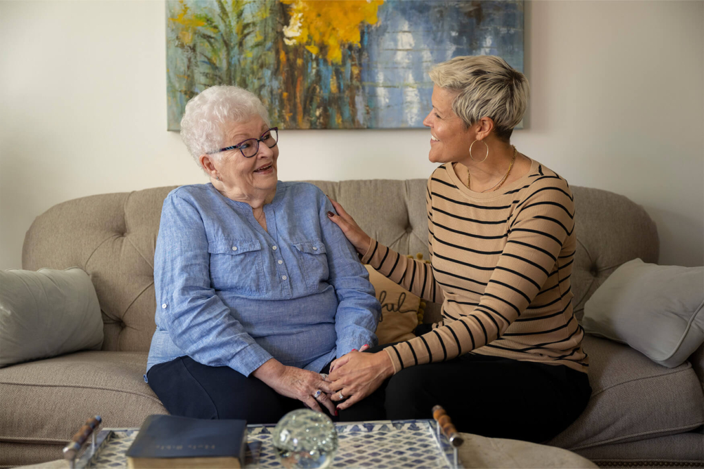 A young woman places her hand on the shoulder of an older woman as they laugh together on a couch inside a 1-bedroom senior apartment