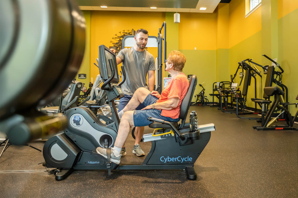 A senior resident sits on a workout machine in the fitness center of a retirement community