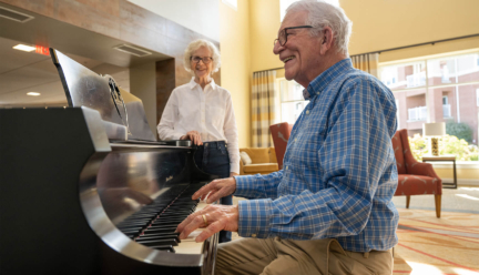 A senior man plays piano in a retirement community while his wife watches