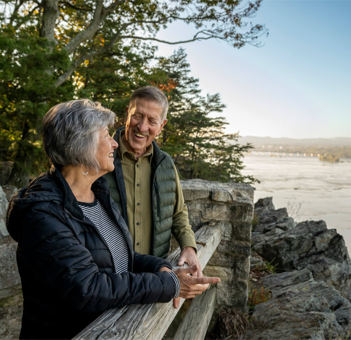 A couple stands at the edge of a lookout overlooking a river and smile at each other