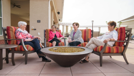 Four women on the senior living pre-registration waitlist chat on a patio, sitting in chairs around a fire pit.
