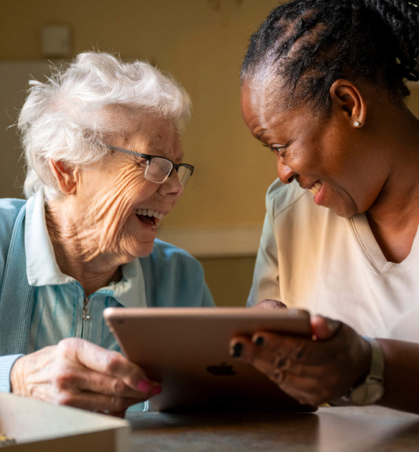 A female nurse holds up a tablet to an older woman as they laugh together