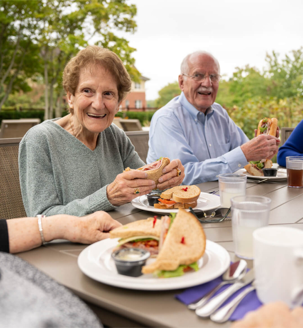Four residents of the Bloom Wellness Woodcrest Villa program eat lunch outside at a table, laughing together