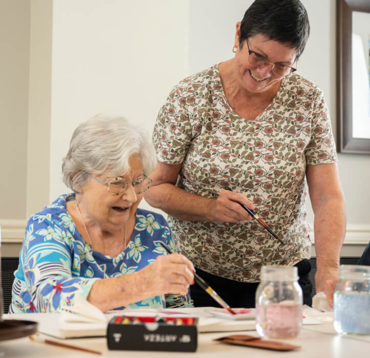 A resident of the Bloom Wellness Woodcrest Villa program teaches an older woman how to paint watercolors