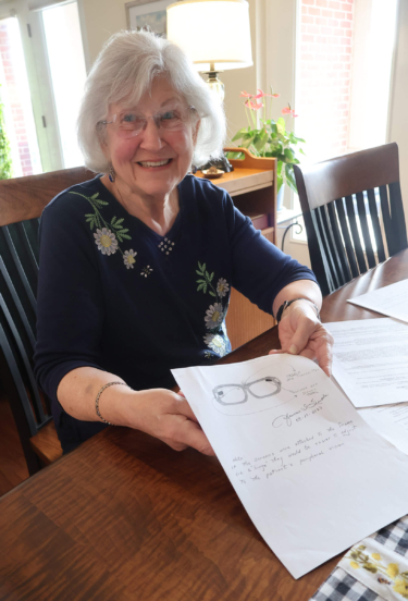 Fran, a resident of a retirement community in Lancaster county, PA, smiles at a table as she holds a paper showing her patented glasses