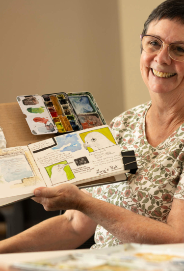 Marge, a resident of a retirement community in Lancaster County, PA, holds up her watercolor book and smiles