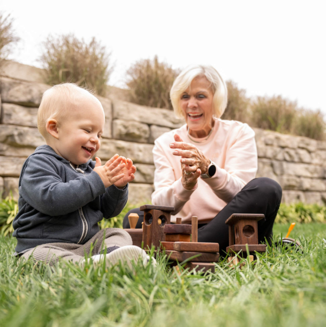 A resident of a retirement community in Lancaster County, PA plays with wooden blocks in the grass with her grandchild. They both clap their hands together and smile
