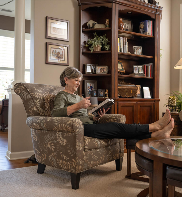 A woman lounges in a chair in her villa, holding a mug and reading a book as she enjoys senior living near Willow Street, PA